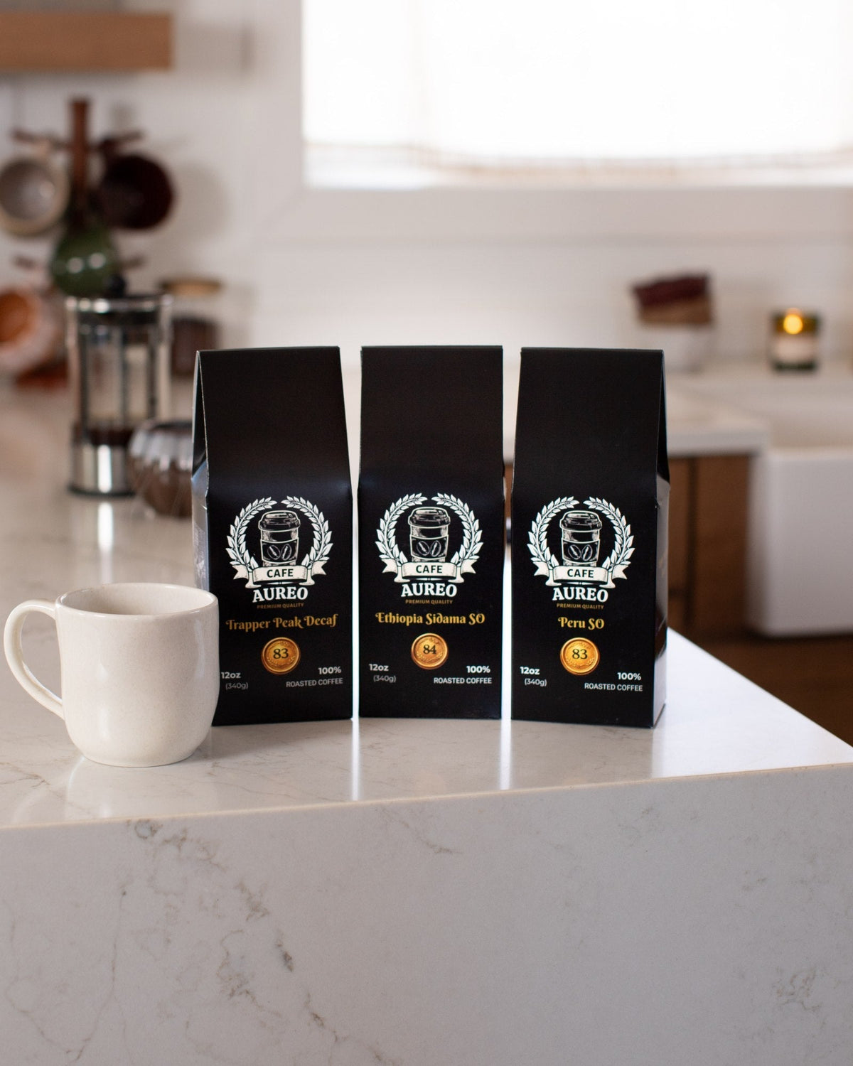 Three AUREO coffees, Trapper Peak Decaf, Ethiopia Sidama SO and Peru SO, beside a mug on a kitchen counter